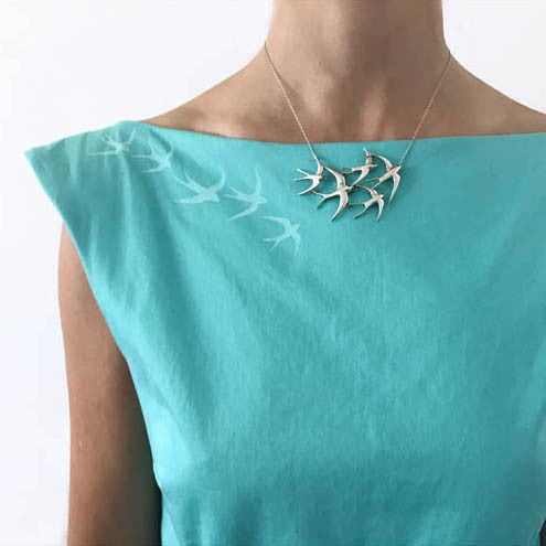 A necklace of a flock of five sterling silver swallow hangs against a turquoise boat neck dress that has silk screened swallows that fly into the necklace.  