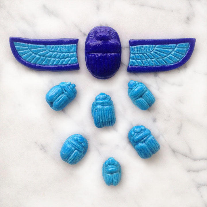 Egyptian faience turquoise and cobalt scarabs and wings sit on a marble surface